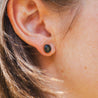 A woman wears black granite round stud earrings made with natural stone. The stone is set in a 14k gold cup.